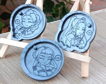 Könnte beinhalten: Drei graue Untersetzer aus Kunstharz mit Cartoon-Illustrationen von Charakteren im Anime-Stil. Jeder Untersetzer hat ein anderes Charakterdesign mit Details wie Frisuren und Kleidung. Die Untersetzer werden auf kleinen Holzstaffeleien ausgestellt.