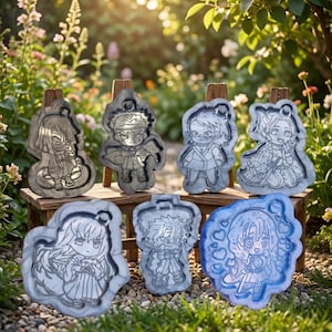 Könnte beinhalten: Eine Sammlung von sieben charakterförmigen Harzformen, jede mit einem einzigartigen Design. Die Formen werden auf einem kleinen Holztisch vor einem Hintergrund aus Grünzeug ausgestellt. Die Formen sind in Grau- und Blautönen gehalten.