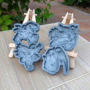 Könnte beinhalten: Vier graue Kunstharzformen mit Cartoon-Charakter-Designs. Die Formen werden auf kleinen Holzstaffeleien präsentiert, die detaillierte Illustrationen verschiedener Kreaturen zeigen. Der Hintergrund umfasst Grün und eine hell geflieste Oberfläche.