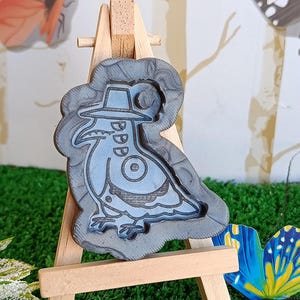 Könnte beinhalten: Ein blau-graues Dekorationsstück in Form eines Vogels mit Hut, ausgestellt auf einer kleinen Holzstaffelei. Der Vogel hat einen langen Schnabel und ist von einem grauen Rand umgeben. Der Hintergrund umfasst Kunstrasen und Schmetterlingsdekorationen.