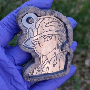 Könnte beinhalten: Ein Holzschlüsselanhänger mit einem lasergravierten Bild eines Comic-Charakters mit Hut. Der Schlüsselanhänger ist braun mit einer dunklen Holzmaserung.