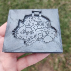 Könnte beinhalten: Ein graues Quadrat mit einem eingravierten Design eines lächelnden Katzenbus-Charakters. Der Katzenbus hat einen detaillierten Buskörper und einen langen, gestreiften Schwanz. Das Design ist in die graue Oberfläche eingraviert.