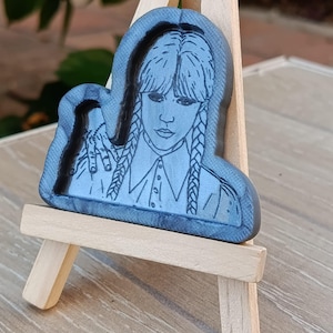 Könnte beinhalten: Eine blau-graue Kunstharzfigur einer Frau mit geflochtenen Zöpfen, die auf einer kleinen Holzstaffelei ausgestellt ist. Die Figur ist ein Porträt mit ernstem Ausdruck, das ein Hemd mit Kragen trägt. Der Hintergrund ist verschwommen und deutet auf eine Umgebung im Freien hin.