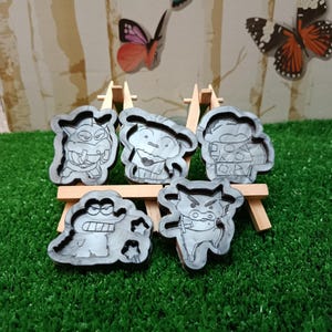 Könnte beinhalten: Fünf graue Cartoon-Charakterformen, die auf kleinen Holzstaffeleien vor einem Hintergrund aus Schmetterlingen und einer grünen Oberfläche ausgestellt sind. Jede Form zeigt ein anderes Charakterdesign mit schwarzen Umrissen und Details.