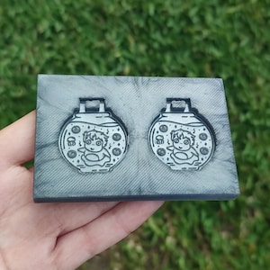 Könnte beinhalten: Zwei graue Harzanhänger mit einer Cartoonfigur in einem Fischglas-Design. Die Anhänger befinden sich auf einer rechteckigen grauen Basis. Der Hintergrund ist verschwommenes grünes Gras.