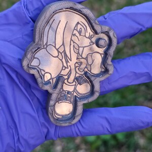 Könnte beinhalten: Ein brauner und grauer Schlüsselanhänger mit einer detaillierten Gravur der Sonic the Hedgehog-Figur Knuckles. Der Schlüsselanhänger hat ein kreisförmiges Loch zur Befestigung und wird in einer behandschuhten Hand gehalten.