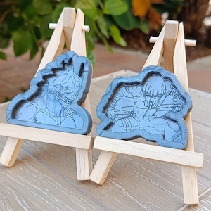 Könnte beinhalten: Zwei blaue Kunstharzfiguren von Charakteren, jeweils auf einer kleinen Holzstaffelei ausgestellt. Die Figuren haben detaillierte Designs, eine zeigt ein Monster und die andere eine Person in einem Anzug. Der Hintergrund ist mit grünem Laub verschwommen.