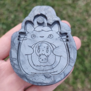 Könnte beinhalten: Ein grauer Resin-Anhänger mit einem Cartoon-Charakter-Design. Der Anhänger hat oben eine Schlaufe zum Aufhängen und zeigt einen Charakter, der einen anderen Charakter hält. Das Design ist in das Harz geschnitzt.