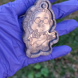 Könnte beinhalten: Ein Holzschlüsselanhänger mit einer Comicfigur, die eine Brille und einen Schnurrbart trägt. Die Figur hält in der einen Hand eine Tasse Kaffee und in der anderen eine Pistole. Der Schlüsselanhänger ist braun und hat eine Holzmaserung.