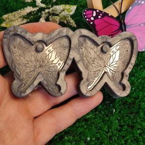 Könnte beinhalten: Zwei schmetterlingsförmige Formen mit detaillierten Flügelmustern. Die Formen sind dunkelgrau mit goldfarbenen Akzenten. Im Hintergrund ist ein rosa und lila Schmetterling zu sehen.