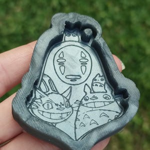 Könnte beinhalten: Eine graue Harzform mit Figuren aus den Zeichentrickfilmen "Chihiros Reise ins Zauberland" und "Mein Nachbar Totoro". Die Form zeigt das gesichtslose Wesen Ohngesicht, Totoro und den Katzenbus. Die Form wird in einer Hand vor einem verschwommenen grünen Hintergrund gehalten.