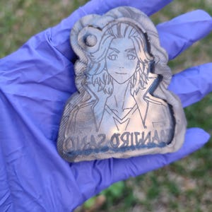 Könnte beinhalten: Ein Schlüsselanhänger aus grauem und schwarzem Harz mit einer Schwarzweiß-Zeichnung einer Person mit langen Haaren. Der Schlüsselanhänger trägt den Text "Onaz Original" unten.