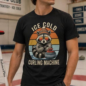 Puede incluir: Camiseta negra con un mapache con gafas de sol y sombrero, sosteniendo una piedra de curling. La camiseta tiene el texto "ICE COLD CURLING MACHINE" en un diseño circular. La imagen también muestra un letrero del club de curling y equipo de curling.