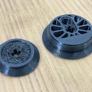 May include: Two black 3D printed circular components. The smaller piece features a textured hexagonal center with a central hole. The larger piece has a raised rim and a complex internal structure with multiple open segments and a central opening.
