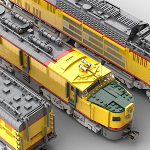 Puede incluir: Una locomotora de tren de juguete amarilla y gris con un frente verde y una franja roja. La locomotora tiene un diseño detallado con múltiples ventanas y una gran parrilla delantera.