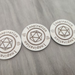 May include: Three wooden round tokens with the word "Inspiration" written around the edge. Each token features a stylized image of a 20-sided die in the centre.