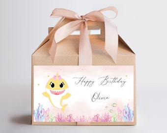 Pode incluir: Uma caixa de presente castanha amarrada com uma fita rosa claro. A caixa tem um rótulo com um desenho de tubarão de desenho animado usando um laço rosa e as palavras "Happy Birthday Olivia". O fundo é um design em aquarela com corais rosa, azuis e roxos.