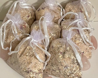 Op de afbeelding: Acht kleine witte organza zakjes gevuld met een mengsel van havervlokken, rozenblaadjes en andere badzouten. De zakjes zijn vastgebonden met wit lint en zijn in een cirkel op een wit oppervlak gerangschikt. Er zijn drie roze hartvormige stenen in de rechterbenedenhoek van de afbeelding.