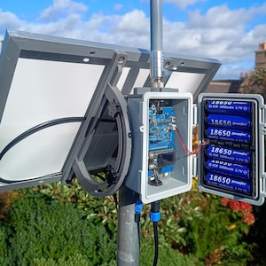 Op de afbeelding: Een zonnepaneelsysteem gemonteerd op een paal, met een grijze behuizing met een printplaat en zes 18650 3,7V batterijen. Het systeem omvat een zonnepaneel en een blauwe en zwarte kabel. De achtergrond toont een blauwe lucht met wolken en groene bladeren.