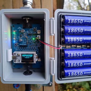 Op de afbeelding: Een open grijze weerbestendige behuizing onthult een elektronische printplaat met een klein display en verschillende blauwe 18650-batterijen. De printplaat heeft verschillende componenten en LED-lampjes. Het display leest "MESHCORE".