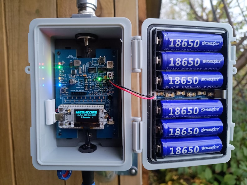 Op de afbeelding: Een open grijze weerbestendige behuizing onthult een elektronische printplaat met een klein display en verschillende blauwe 18650-batterijen. De printplaat heeft verschillende componenten en LED-lampjes. Het display leest "MESHCORE".