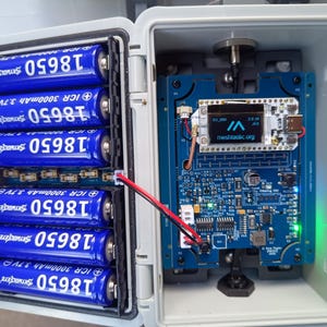 Op de afbeelding: Een open, witte plastic behuizing onthult een elektronisch apparaat met een blauwe printplaat en een klein scherm met "meshtastic.org". Zes blauwe 18650-batterijen zijn links zichtbaar. Een rood-zwarte draad verbindt de batterijen met de printplaat.