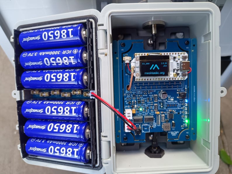 Op de afbeelding: Een open, witte plastic behuizing onthult een elektronisch apparaat met een blauwe printplaat en een klein scherm met "meshtastic.org". Zes blauwe 18650-batterijen zijn links zichtbaar. Een rood-zwarte draad verbindt de batterijen met de printplaat.