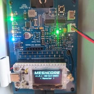Op de afbeelding: Een close-up van een blauwe printplaat met een scherm dat "MESHCORE v1.9.1 (02-Oct-2025) < Repeater >" weergeeft. De printplaat bevat diverse elektronische componenten, waaronder LED's en weerstanden, en is geplaatst in een witte behuizing.