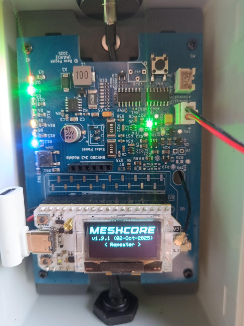 Op de afbeelding: Een close-up van een blauwe printplaat met een scherm dat "MESHCORE v1.9.1 (02-Oct-2025) < Repeater >" weergeeft. De printplaat bevat diverse elektronische componenten, waaronder LED's en weerstanden, en is geplaatst in een witte behuizing.