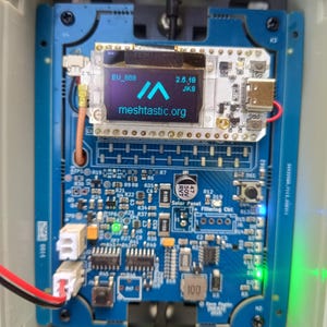 Op de afbeelding: Een blauwe printplaat met diverse elektronische componenten, waaronder een klein scherm met de tekst "meshtastic.org". De printplaat heeft meerdere connectoren, een USB-poort en de tekst "EU_868" en "2.5.18 JKS". Het is gemonteerd in een grijze behuizing.