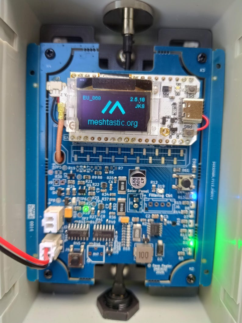 Op de afbeelding: Een blauwe printplaat met diverse elektronische componenten, waaronder een klein scherm met de tekst "meshtastic.org". De printplaat heeft meerdere connectoren, een USB-poort en de tekst "EU_868" en "2.5.18 JKS". Het is gemonteerd in een grijze behuizing.