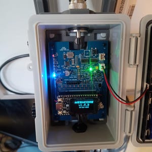 Op de afbeelding: Een grijze, rechthoekige behuizing bevat een blauwe printplaat met diverse elektronische componenten, waaronder een klein scherm met de tekst "MESHCORE v1.9.0". De behuizing heeft een zilveren antenne en is gemonteerd op een wit oppervlak. Verschillende LED-lampjes zijn verlicht.