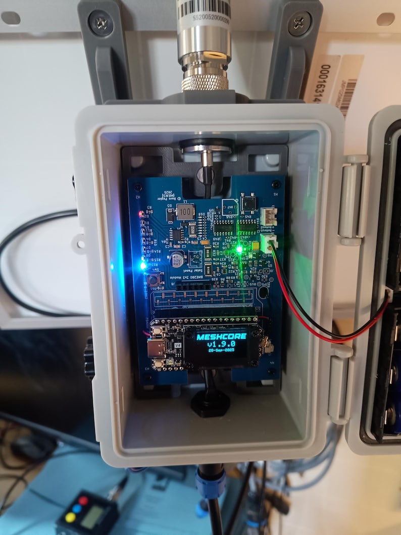 Op de afbeelding: Een grijze, rechthoekige behuizing bevat een blauwe printplaat met diverse elektronische componenten, waaronder een klein scherm met de tekst "MESHCORE v1.9.0". De behuizing heeft een zilveren antenne en is gemonteerd op een wit oppervlak. Verschillende LED-lampjes zijn verlicht.