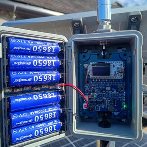 Op de afbeelding: Een open grijze weerbestendige behuizing met blauwe 18650-batterijen en een printplaat. De batterijen zijn gemarkeerd met "ICR 3000mAh 3.7V". Een rode en zwarte draad verbindt de batterijen met de printplaat. Een zonnepaneel is op de achtergrond.