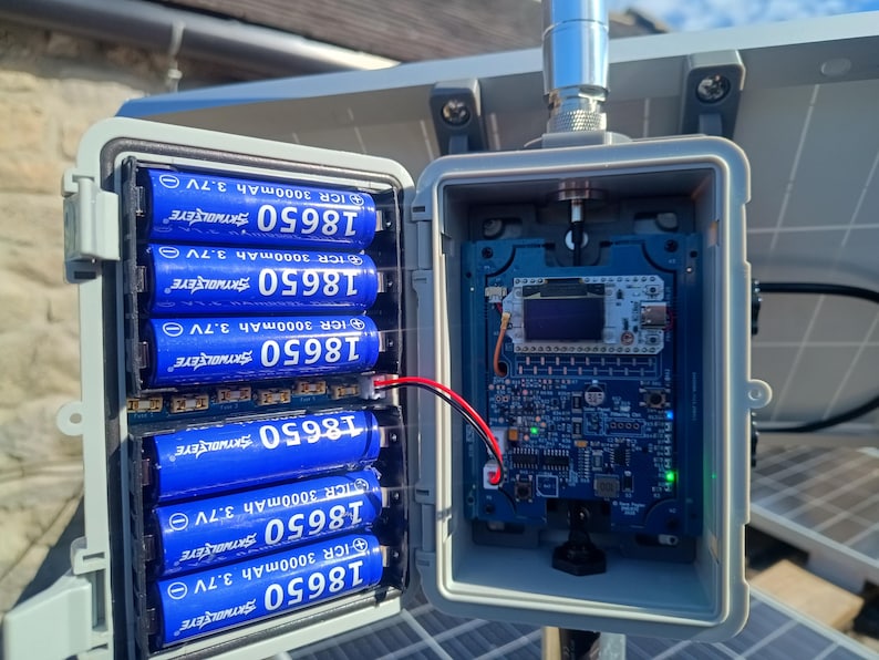 Op de afbeelding: Een open grijze weerbestendige behuizing met blauwe 18650-batterijen en een printplaat. De batterijen zijn gemarkeerd met "ICR 3000mAh 3.7V". Een rode en zwarte draad verbindt de batterijen met de printplaat. Een zonnepaneel is op de achtergrond.