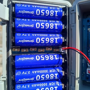 Op de afbeelding: Een open grijze plastic behuizing toont zeven blauwe 18650 oplaadbare batterijen. Elke batterij is voorzien van witte tekst, waaronder "SKYWOLFEYE", "ICR 3000mAh 3.7V" en "18650". Draden komen uit het batterijcompartiment.