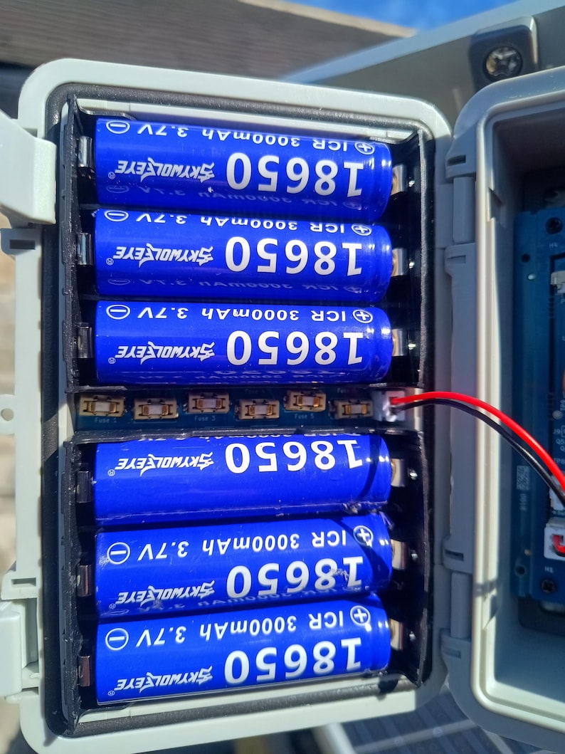 Op de afbeelding: Een open grijze plastic behuizing toont zeven blauwe 18650 oplaadbare batterijen. Elke batterij is voorzien van witte tekst, waaronder "SKYWOLFEYE", "ICR 3000mAh 3.7V" en "18650". Draden komen uit het batterijcompartiment.