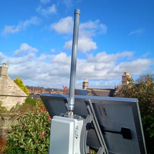 Op de afbeelding: Een zonnepaneelopstelling gemonteerd op een paal, met twee grijze zonnepanelen, een witte doos en een grijze antenne. De achtergrond toont een blauwe lucht met wolken en een woonwijk.