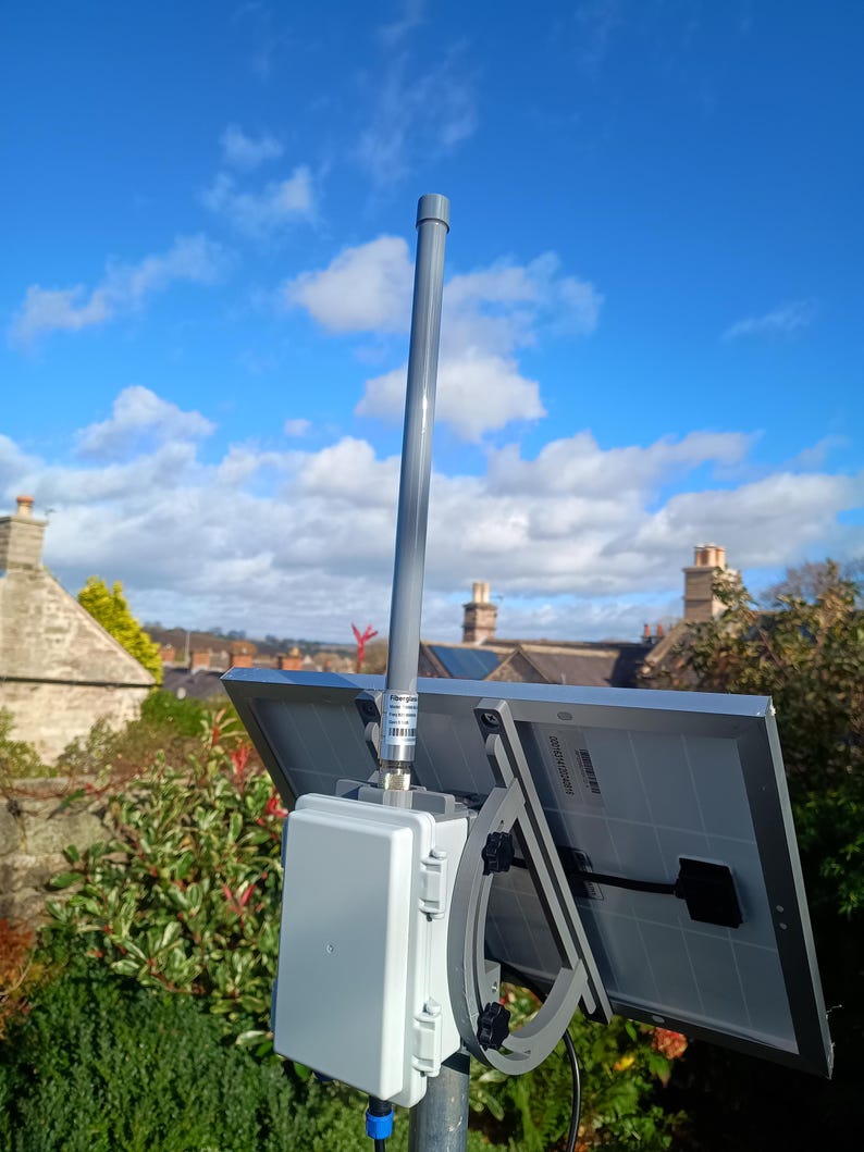 Op de afbeelding: Een zonnepaneelopstelling gemonteerd op een paal, met twee grijze zonnepanelen, een witte doos en een grijze antenne. De achtergrond toont een blauwe lucht met wolken en een woonwijk.