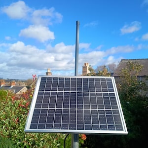 Op de afbeelding: Een zonnepaneel met een zilveren frame is gemonteerd op een grijze paal. Het paneel is zwart met een rasterpatroon. De achtergrond toont een blauwe lucht met wolken, groene struiken en huizen.
