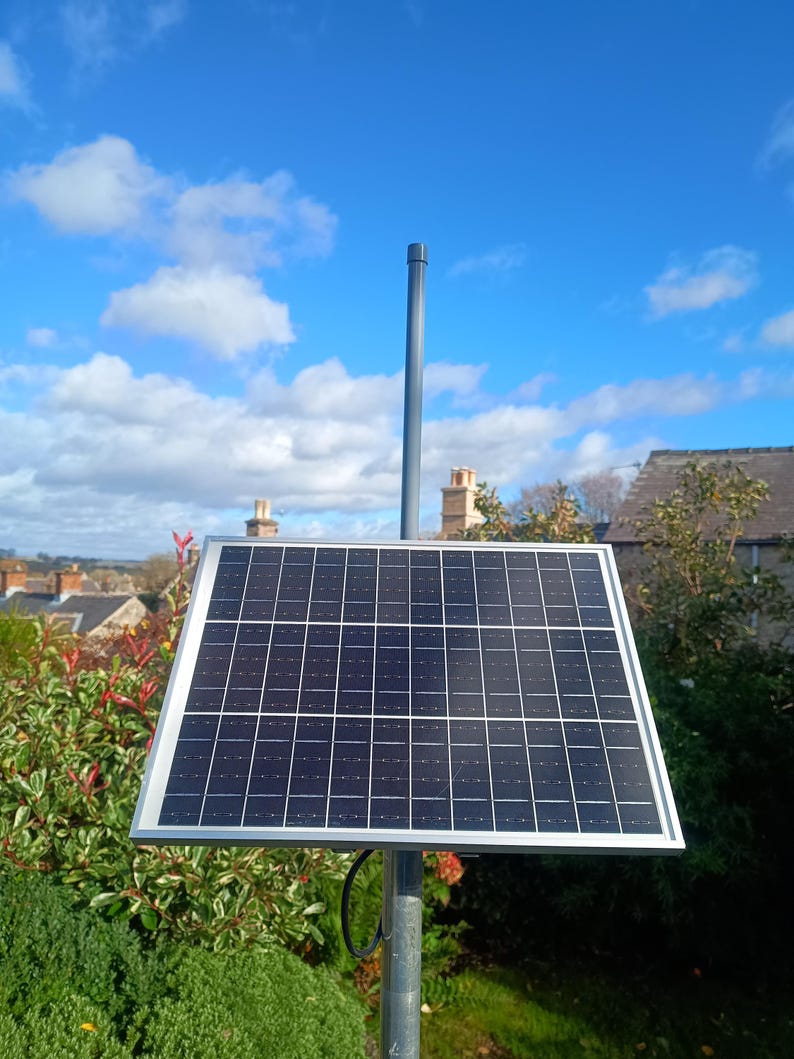 Op de afbeelding: Een zonnepaneel met een zilveren frame is gemonteerd op een grijze paal. Het paneel is zwart met een rasterpatroon. De achtergrond toont een blauwe lucht met wolken, groene struiken en huizen.