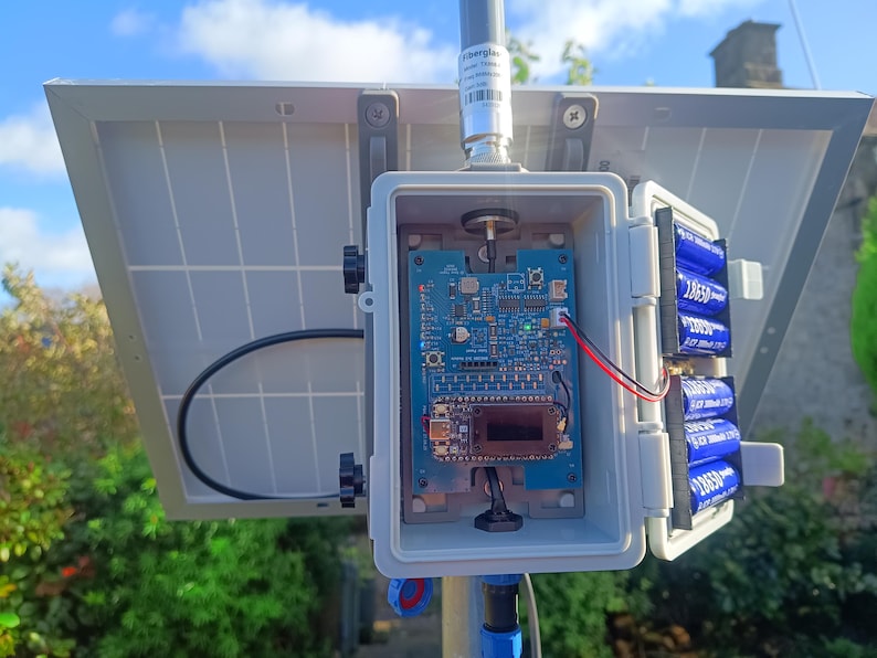 Op de afbeelding: Een zonnepaneel met een printplaat in een grijze doos. De doos is op een paal gemonteerd en heeft een batterijcompartiment met vier blauwe 18650 batterijen. Een zwarte kabel en een antenne zijn ook zichtbaar.