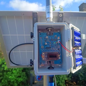 Op de afbeelding: Een zonnepaneelsysteem met een grijze behuizing, een printplaat en blauwe batterijen. Het systeem is op een paal gemonteerd. De printplaat bevat diverse elektronische componenten.