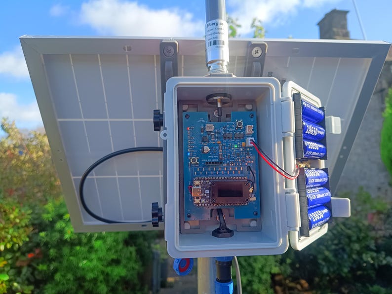 Op de afbeelding: Een zonnepaneelsysteem met een grijze behuizing, een printplaat en blauwe batterijen. Het systeem is op een paal gemonteerd. De printplaat bevat diverse elektronische componenten.