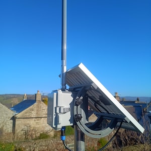 Może przedstawiać: Panel słoneczny zamontowany na szarym słupie, skierowany w stronę słońca. Panel jest przymocowany do szarej skrzynki z czarnym kablem. Tło przedstawia czyste błękitne niebo i widok na miasto.