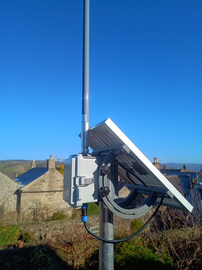 Op de afbeelding: Een zonnepaneel gemonteerd op een grijze paal, gericht op de zon. Het paneel is bevestigd aan een grijze doos met een zwarte kabel. De achtergrond toont een heldere blauwe lucht en een uitzicht op een stad.