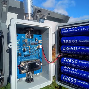 Op de afbeelding: Een grijze buitenbehuizing met een blauwe printplaat, een klein scherm en verschillende blauwe 18650-batterijen. De behuizing is gemonteerd op een paal met een blauwe lucht op de achtergrond.