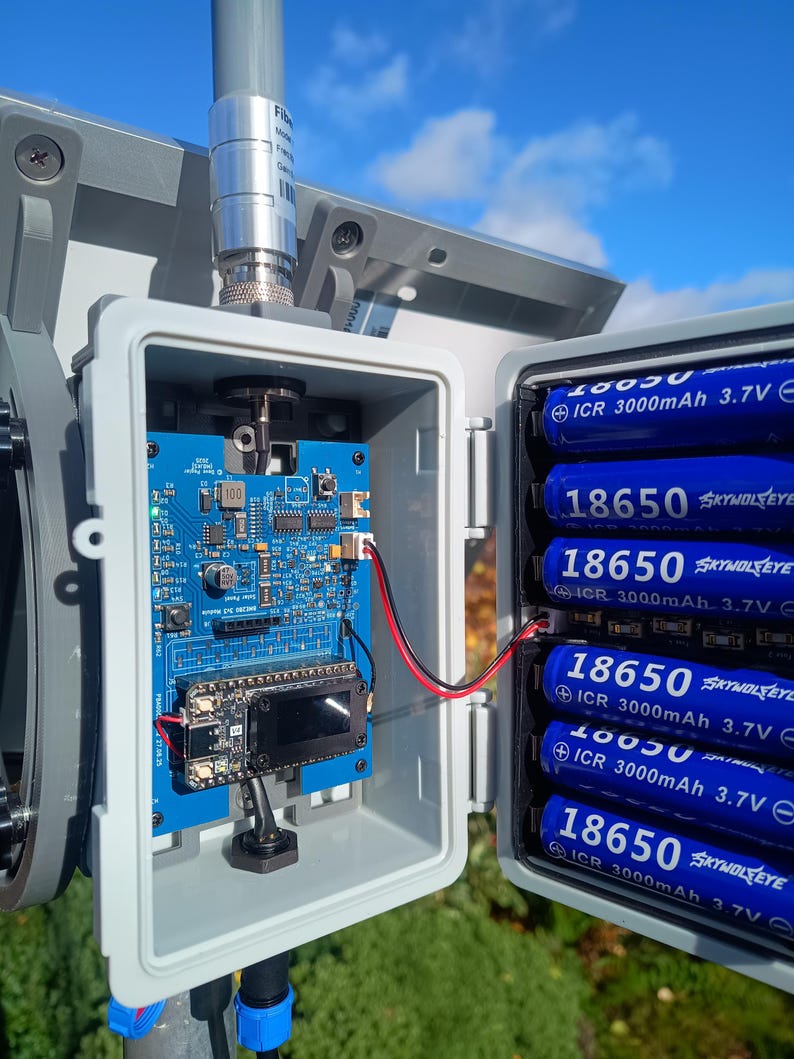 Op de afbeelding: Een grijze buitenbehuizing met een blauwe printplaat, een klein scherm en verschillende blauwe 18650-batterijen. De behuizing is gemonteerd op een paal met een blauwe lucht op de achtergrond.