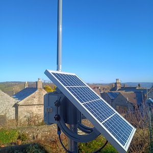 Op de afbeelding: Een zonnepaneel gemonteerd op een grijze paal, gericht op de zon. Het paneel is rechthoekig met een raster van donkerblauwe vierkanten. Een grijze doos en een gebogen beugel ondersteunen het paneel. De achtergrond toont een heldere blauwe lucht en een ver dorp.
