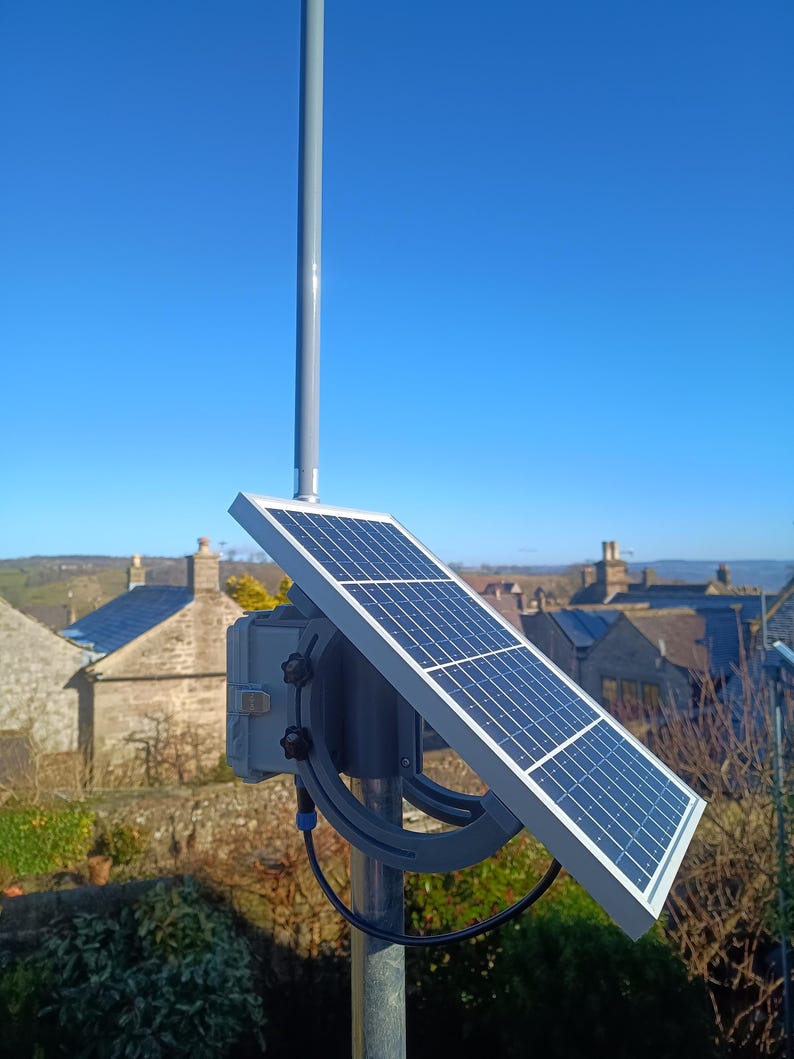 Op de afbeelding: Een zonnepaneel gemonteerd op een grijze paal, gericht op de zon. Het paneel is rechthoekig met een raster van donkerblauwe vierkanten. Een grijze doos en een gebogen beugel ondersteunen het paneel. De achtergrond toont een heldere blauwe lucht en een ver dorp.